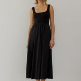 A model wearing a black maxi dress with a knit square neckline top and a woven full skirt. The dress has side pockets, is sleeveless, and reaches down to the ankles.