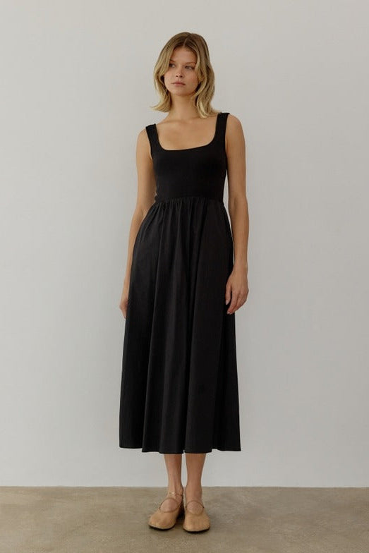 A model wearing a black maxi dress with a knit square neckline top and a woven full skirt. The dress has side pockets, is sleeveless, and reaches down to the ankles.