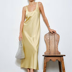 Woman in a light yellow dress standing next to a wooden chair against a white wall.