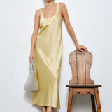 Woman in a light yellow dress standing next to a wooden chair against a white wall.