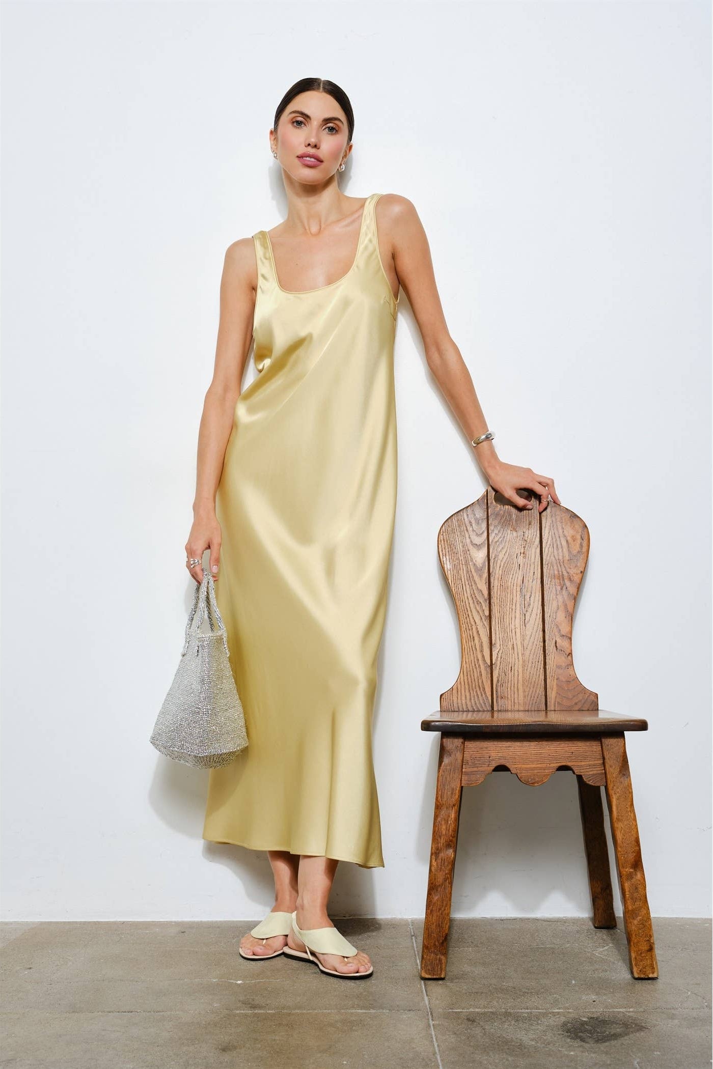 Woman in a light yellow dress standing next to a wooden chair against a white wall.